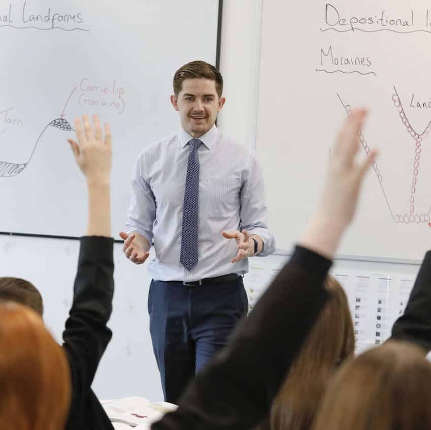 A teacher delivers a lesson to a class at Laurus Ryecroft. Students in the foreground have their arms raised to answer a question.